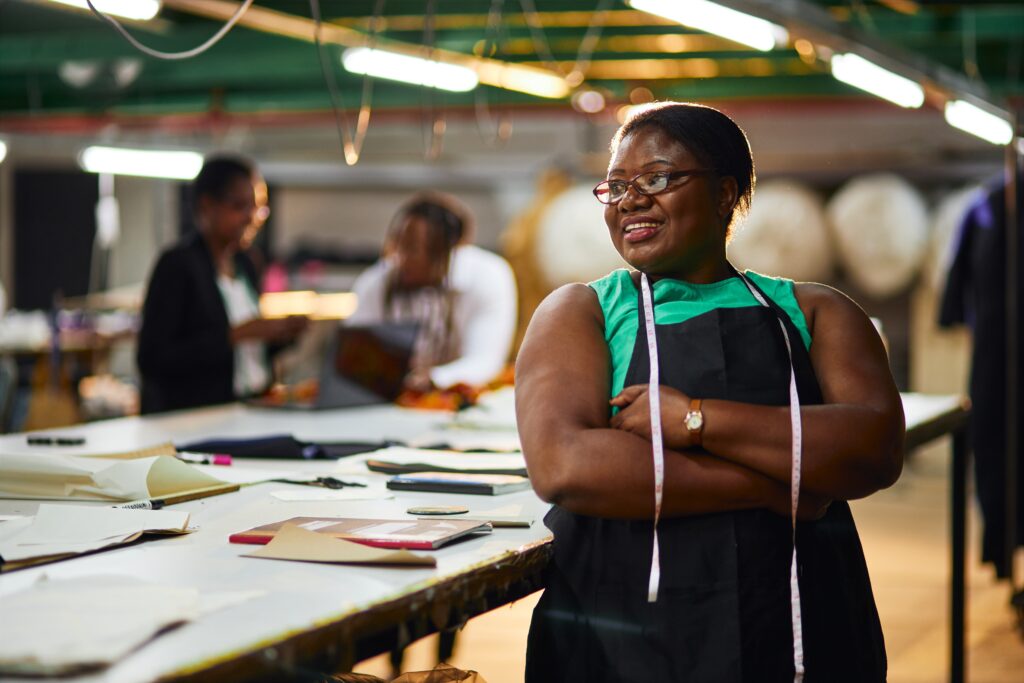 African,Female,Business,Woman,Textile,Industry,Wearing,A,Green,Shirt Eine dunkelhäutige Frau steht selbstbewusst mit einem Maßband umhängend im Raum